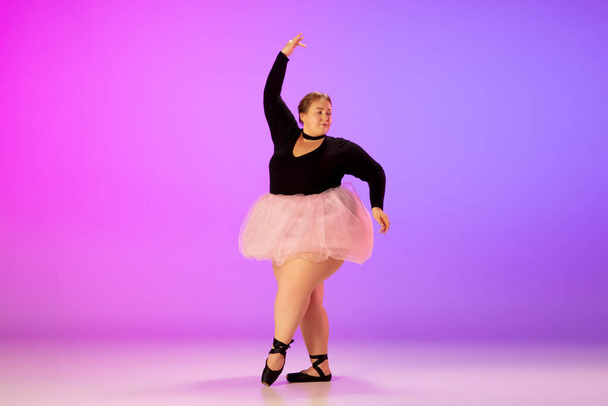 Beautiful caucasian plus size model practicing ballet dance on gradient purple-pink studio background in neon light - Photo, image