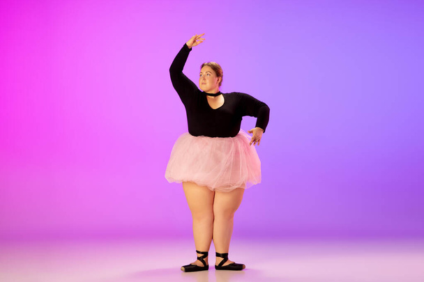 Beautiful caucasian plus size model practicing ballet dance on gradient purple-pink studio background in neon light - Фото, изображение