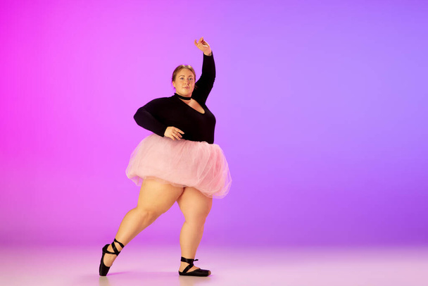 Beautiful caucasian plus size model practicing ballet dance on gradient purple-pink studio background in neon light - Фото, изображение