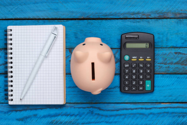 Malacka bank jegyzetfüzettel és számológéppel, kék fa háttérrel. Felülnézet - Fotó, kép