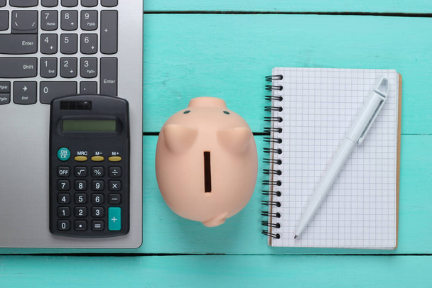 Piggy Bank Met Laptop, Calculator, notitieboekje op blauwe houten achtergrond. Maak geld online of internet business concepten. Bovenaanzicht. Vlakke plaat - Foto, afbeelding