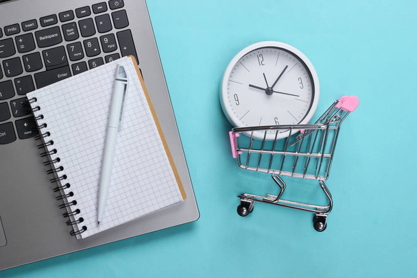 Temps d'achat en ligne. Clavier d'ordinateur avec horloge et chariot supermarché, liste d'achats sur fond bleu. Vue du dessus - Photo, image