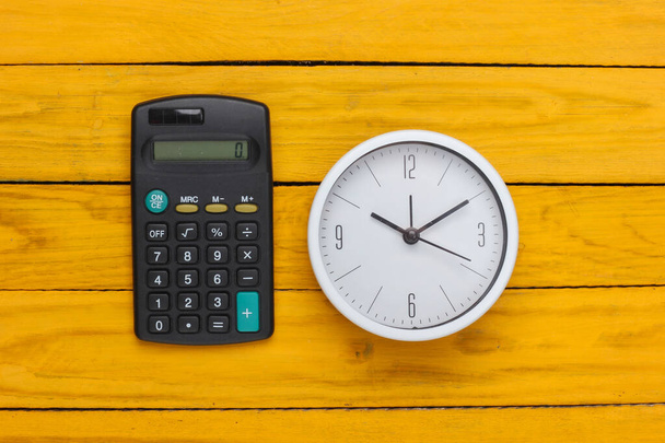 Biały zegar i kalkulator na żółtym drewnianym tle. Minimalistyczne ujęcie studyjne. Widok z góry - Zdjęcie, obraz