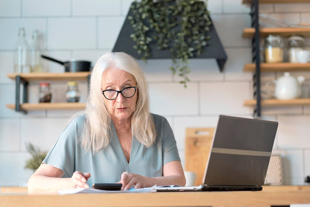 彼女の台所でウェブサーフィンのためのラップトップを使用しているシニア女性。高齢者の雇用、社会保障の概念。自宅のオフィスでノートパソコンをタイプする仕事に座っている熟女. - 写真・画像