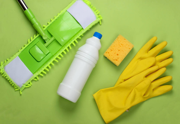 Чистящие средства. Пластиковая швабра, перчатки, бутылка моющего средства, губка на зеленом фоне. Дезинфекция и уборка в доме. Вид сверху - Фото, изображение