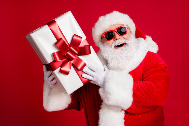 Photo of pensioner old man white beard hold present box ribbon bow-knot curious interested wonder what inside wear santa costume sunglass gloves headwear isolated red color background - Φωτογραφία, εικόνα