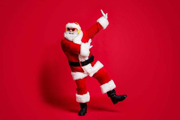 Feel young again. Full length photo of retired old man grey beard raise hands partying have fun wear santa costume sunglass headwear leather black boots isolated red color background - Foto, immagini