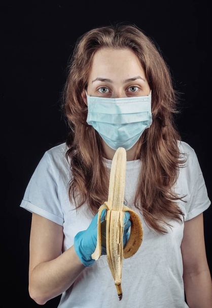 Γυναίκα με προστατευτική ιατρική μάσκα και γάντια προσπαθεί να φάει μια μπανάνα σε μαύρο φόντο - Φωτογραφία, εικόνα