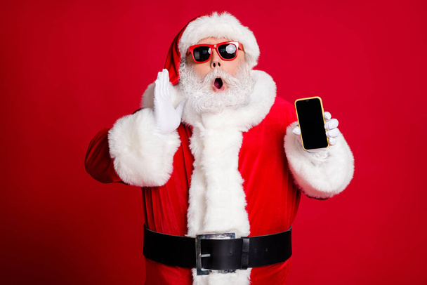 Photo of pensioner old man grey beard hold telephone empty space hand open mouth yell sale information wear x-mas santa costume gloves coat belt sunglass cap isolated red color background - Zdjęcie, obraz