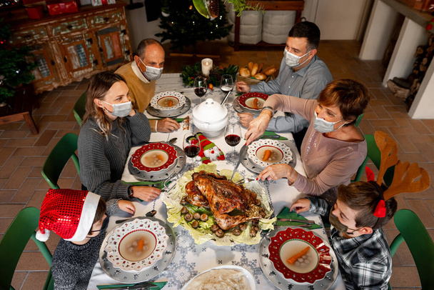 Zenith szög a család gyűlt össze az asztal körül a karácsonyi hálaadási vacsora 2020-ban. A családtagok sebészeti maszkot viselnek, hogy megfeleljenek az új egészségügyi előírásoknak. Békés karantén, lezárás. - Fotó, kép