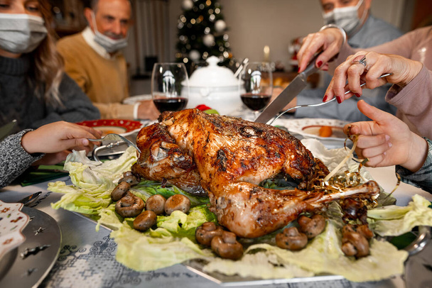 CU of a woman hands with a knife cutting a roast turkey on Christmas Thanksgiving dinner. The members of the family are wearing protective face mask. Covid outbreak, coronavirus pandemic, lockdown. - 写真・画像