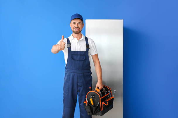 Arbeiter des Reparaturdienstes zeigt Daumen nach oben in der Nähe des Kühlschranks auf farbigem Hintergrund - Foto, Bild