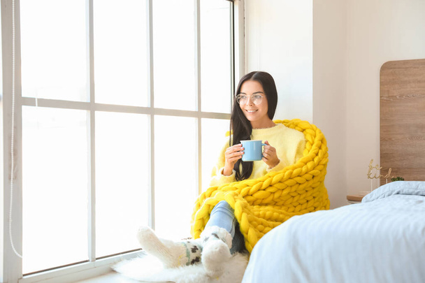Bella giovane donna avvolta in caldo plaid bere tè vicino alla finestra - Foto, immagini
