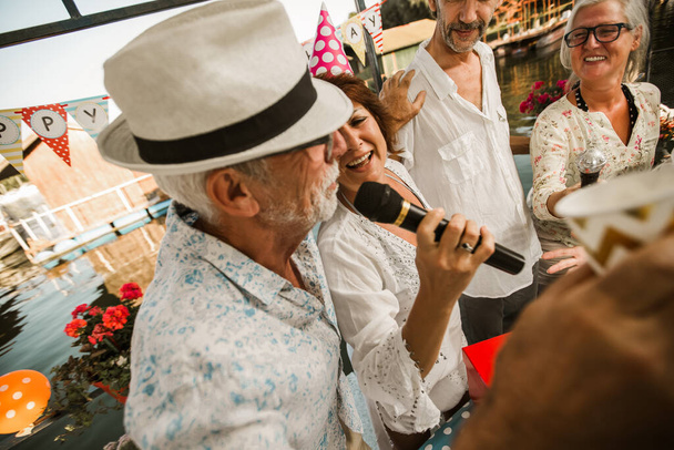 Oudere mensen vieren hun verjaardag in het huisje aan de rivier en zingen karaoke. - Foto, afbeelding