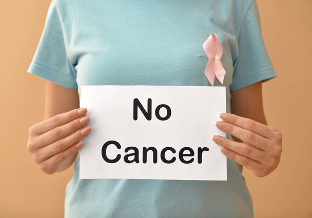 Woman with pink ribbon holding paper sheet with text NO CANCER on color background - Foto, Imagen