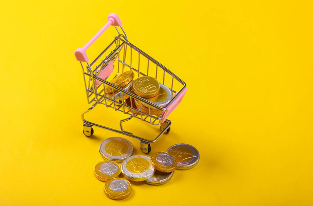 Shopping theme. Mini Supermarket trolley with coins on yellow background. - Фото, зображення