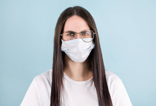 Close-up portrait of cute a girl wear white shirt protection flu cold facial mask isolated over blue pastel background with misted glasses.. - Φωτογραφία, εικόνα