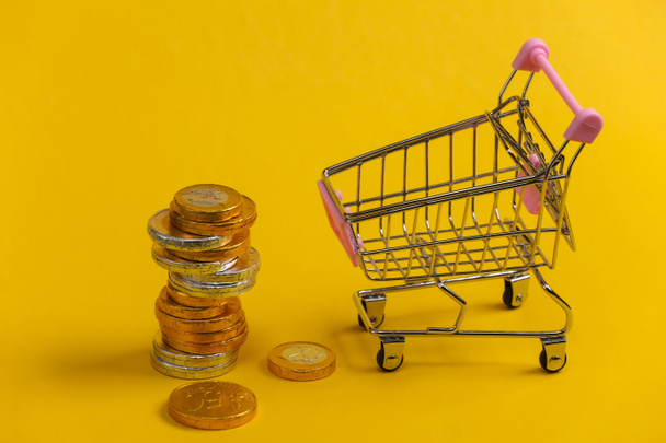 Winkelthema. Mini Supermarkt trolley met munten op gele achtergrond. - Foto, afbeelding