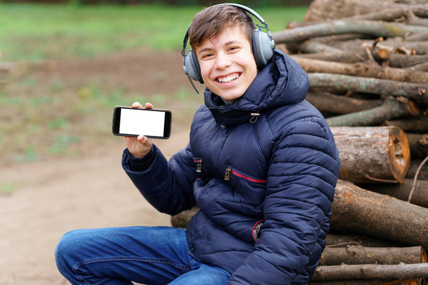 teenager listening to music by headphones, relaxing in autumn city park, shows the phone screen and sitting on a log - Photo, Image