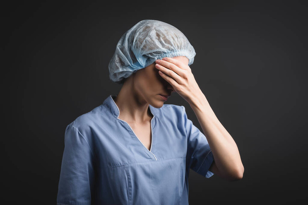 Enfermera cansada en gorra médica cubriendo ojos aislados en gris oscuro - Foto, Imagen