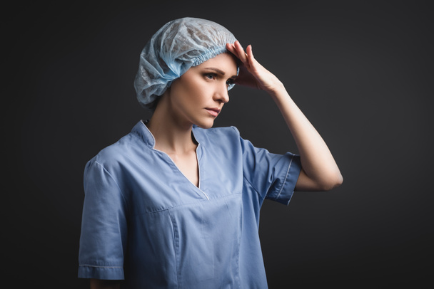 tired nurse in medical cap looking away isolated on dark grey - Foto, imagen