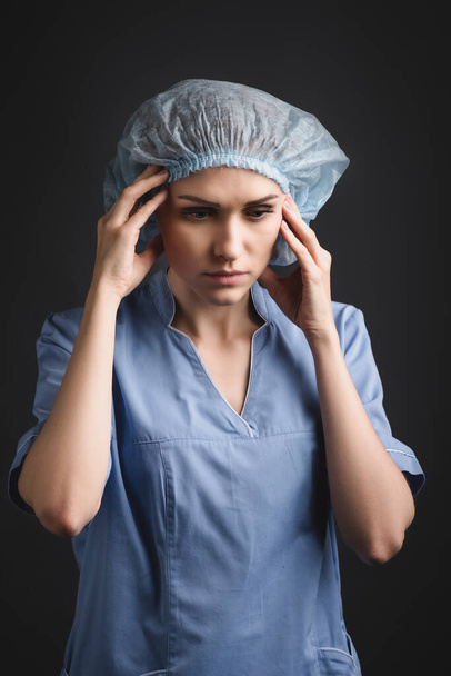 enfermera cansada en gorra médica que sufre de migraña aislada en gris oscuro - Foto, Imagen