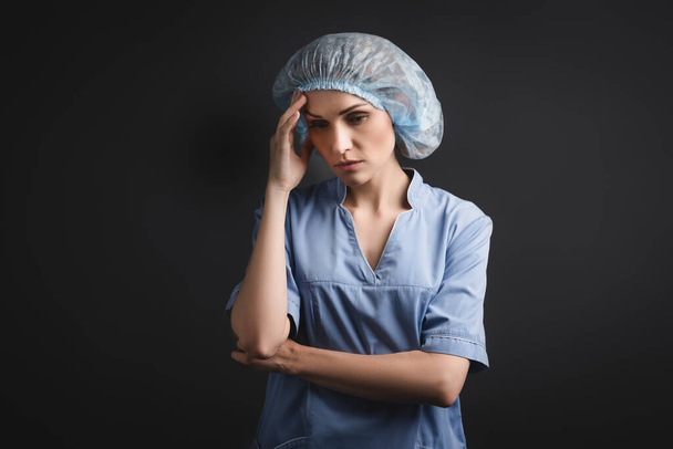 enfermera en gorra médica azul con dolor de cabeza aislado en gris oscuro - Foto, Imagen