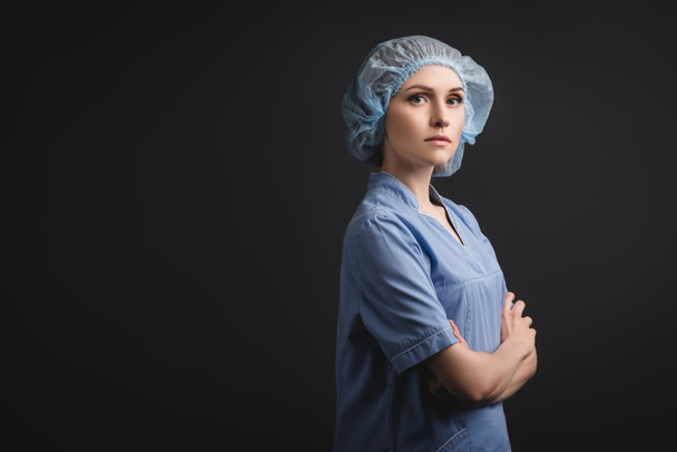 enfermera en gorra médica de pie con brazos cruzados aislados en gris oscuro - Foto, Imagen