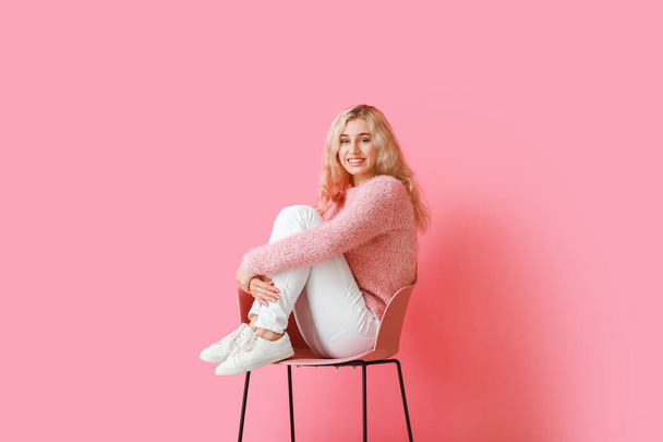 Mooie jonge vrouw in warme kleren zitten op stoel tegen kleur achtergrond - Foto, afbeelding