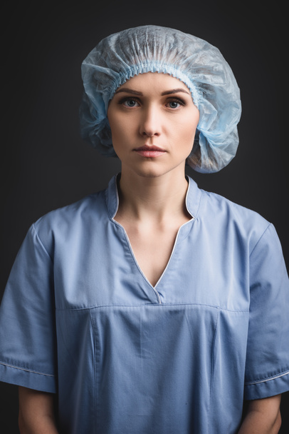 zdravotní sestra v modré lékařské čepice při pohledu na kameru izolované na tmavě šedé - Fotografie, Obrázek