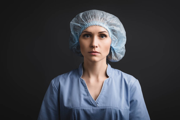 enfermera seria con gorra médica azul mirando a la cámara aislada en gris oscuro - Foto, Imagen