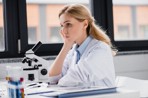 trieste wetenschapper leunend op hand in de buurt van microscoop in lab - Foto, afbeelding