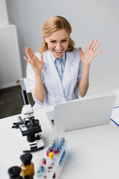Χαρούμενος επιστήμονας με λευκό παλτό κοιτάζοντας το φορητό υπολογιστή κοντά στο μικροσκόπιο στο γραφείο - Φωτογραφία, εικόνα