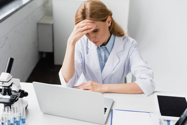 upset scientist in white coat covering face near laptop and microscope on desk - Photo, Image