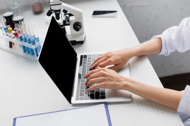 обрізаний вид вченого, що друкує на ноутбуці з порожнім екраном біля мікроскопа на столі
 - Фото, зображення