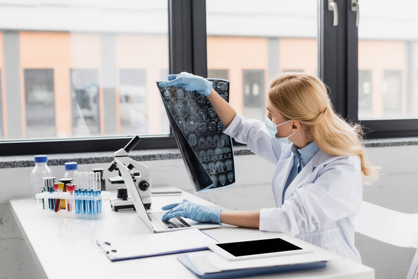 ученый в медицинской маске смотрит на рентген рядом с устройствами на столе - Фото, изображение
