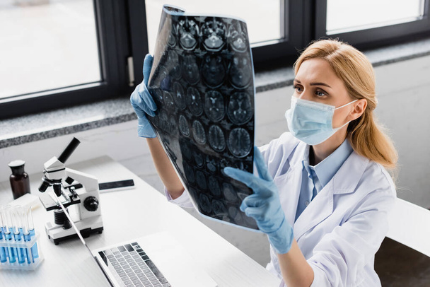 Wissenschaftler in medizinischer Maske betrachtet röntgennahe Geräte und Mikroskop auf dem Schreibtisch - Foto, Bild