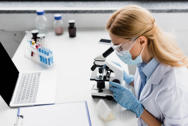 ученый в медицинской маске смотрит через микроскоп на столе - Фото, изображение