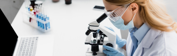 naukowiec w masce medycznej patrząc przez mikroskop na biurku, baner - Zdjęcie, obraz