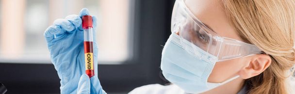 naukowiec w masce medycznej i goglach patrząc na probówkę z jaskrawym napisem, baner - Zdjęcie, obraz