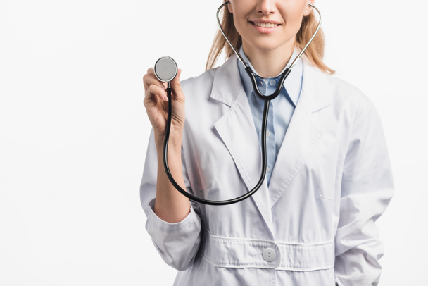 bijgesneden weergave van gelukkige verpleegster in witte vacht bedrijf stethoscoop geïsoleerd op wit - Foto, afbeelding