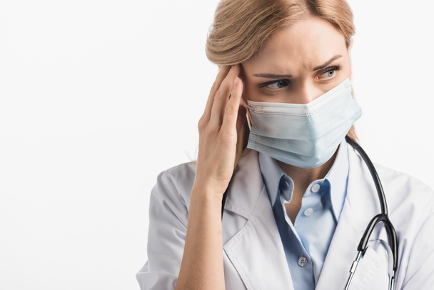 enfermera en máscara médica y bata blanca que sufre de migraña aislada en blanco - Foto, Imagen