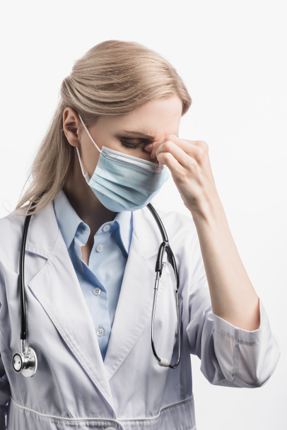 Müde Krankenschwester in medizinischer Maske und weißem Kittel mit Kopfschmerzen isoliert auf weißem Untergrund - Foto, Bild