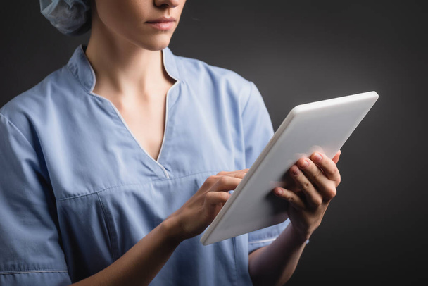 bijgesneden weergave van verpleegkundige in uniform met behulp van digitale tablet geïsoleerd op donkergrijs - Foto, afbeelding