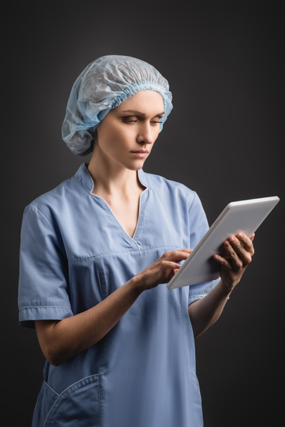 infirmière en casquette médicale et uniforme à l'aide d'une tablette numérique isolée sur gris foncé - Photo, image