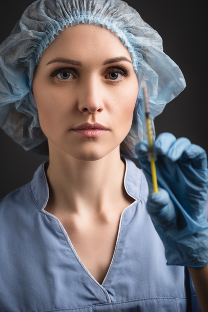 enfermera con gorra médica y jeringa uniforme con vacuna en primer plano borrosa aislada en gris oscuro - Foto, Imagen