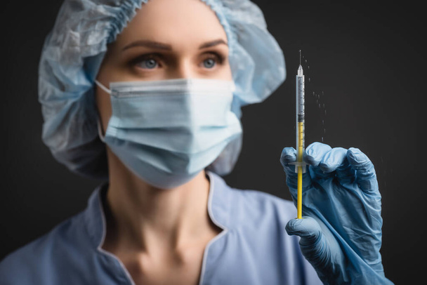 jeringa con salpicadura de vacuna en la mano de la enfermera con máscara médica sobre fondo borroso aislado sobre gris oscuro - Foto, Imagen