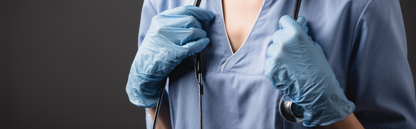 vista recortada de la enfermera en guantes de látex ajustando estetoscopio aislado en gris oscuro  - Foto, imagen