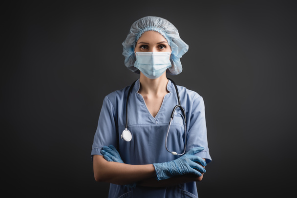 nurse in medical mask and latex gloves standing with crossed arms isolated on dark grey  - Photo, Image
