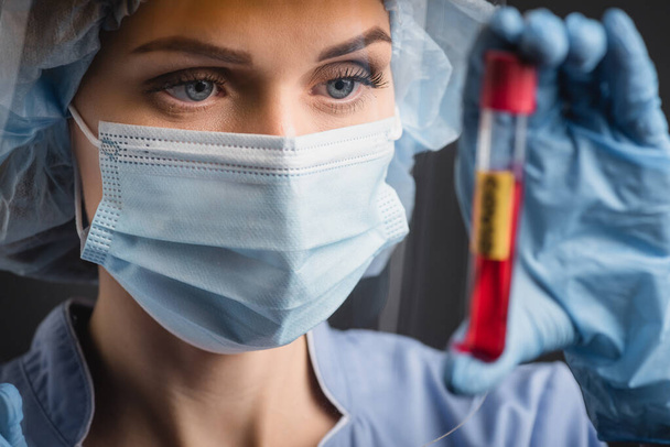 enfermera en máscara médica sosteniendo tubo de ensayo en primer plano borroso aislado en gris oscuro - Foto, Imagen